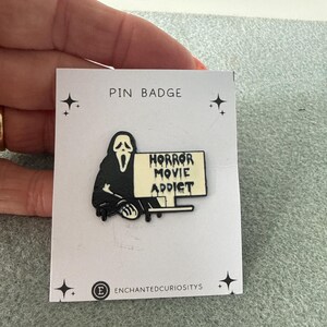 May include: A black and white enamel pin badge featuring a cloaked figure with a white mask holding a sign that reads HORROR MOVIE ADDICT in a dripping font. The pin is attached to a white display card labelled PIN BADGE and ENCHANTEDCURIOSITYS.