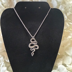 May include: Silver snake pendant necklace. The pendant, a detailed snake design, is coiled and textured. The necklace is displayed on a black stand against a backdrop of white flowers.