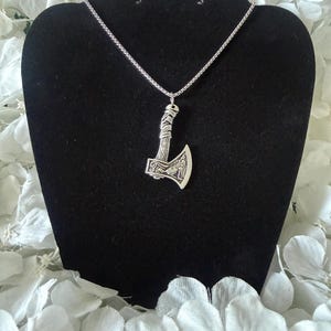 May include: A silver axe pendant necklace. The pendant, with detailed carvings and a wrapped handle, hangs from a fine chain. The necklace is displayed on a black stand, set against a backdrop of white flowers. The axe pendant is a striking piece of jewellery.