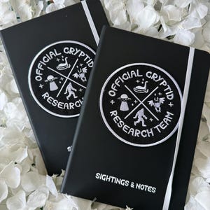 May include: Two black notebooks with white elastic bands, each featuring a circular embroidered patch that reads OFFICIAL CRYPTID RESEARCH TEAM. The patch displays icons of a creature, a UFO, and a moth-like figure. The text SIGHTINGS & NOTES is printed below.