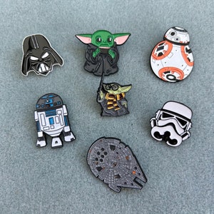May include: A selection of enamel pins depicting characters from a well-known sci-fi series. The pins showcase Darth Vader, Grogu, BB-8, R2-D2, a Stormtrooper, and the Millennium Falcon, in various colours.