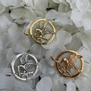 May include: Three Mockingjay pins, a symbol from the Hunger Games, in gold and silver finishes. The pins feature a stylised mockingjay bird with its wings spread, perched on an arrow.