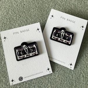 May include: Two enamel pins shaped like cassette tapes, each featuring a white ghost-like mask in the centre. The text on the pins reads "What's your favourite scary movie?" The pins are attached to white backing cards labelled "PIN BADGE" and "ENCHANTEDCURIOSITYS".