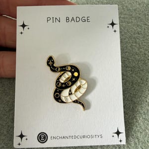 May include: A gold-toned metal pin badge featuring a coiled snake design with black and white enamel detailing, moon phases, and small stars. The pin is attached to a white display card labelled PIN BADGE and ENCHANTEDCURIOSITYS with star accents.