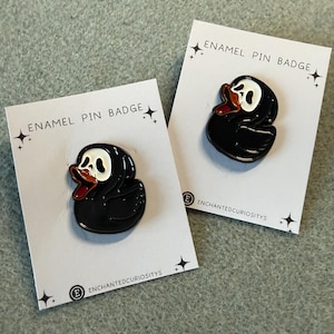 May include: Two enamel pin badges depicting a black duck with a white face and a red beak. The pins are presented on white cards, with the text "ENAMEL PIN BADGE" and the "EnchantedCuriositys" logo.