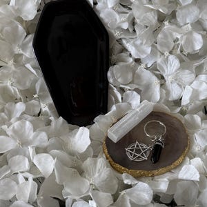May include: A black coffin-shaped tray, a white rectangular selenite crystal, and a silver-toned keyring featuring a pentagram charm and a black crystal point, all resting on a circular brown stone coaster with a metallic gold-coloured edge.
