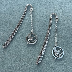 May include: Two silver metal bookmarks with a decorative pattern. Each bookmark has a chain attached to a round pendant featuring a bird with outstretched wings and arrows. The bookmarks are displayed on a blue-grey surface.