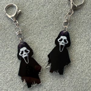 May include: Two keyrings featuring a black figure with a white mask, resembling a ghost. Each keyring has a silver clasp and chain. The keyrings are likely made of acrylic or a similar material.
