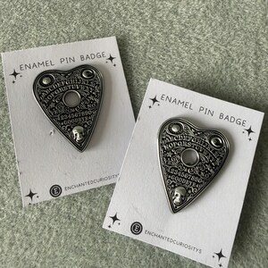 May include: Two silver enamel pin badges shaped like a heart-shaped planchette, with the text "ENAMEL PIN BADGE" at the top. The planchette features the alphabet, numbers, and the words "YES", "NO", and "GOODBYE". The brand name "ENCHANTEDCURIOSITYS" is at the bottom.