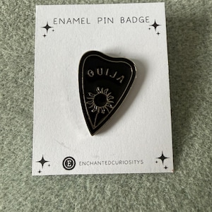 May include: A black enamel planchette-shaped pin badge with the word "OUJIA" in white. The pin has a sun design and is on a white card. The card also features the text "ENAMEL PIN BADGE" and "ENCHANTEDCURIOSITYS".