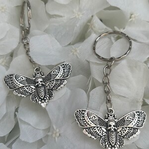May include: Two silver keychains featuring a detailed, ornate moth design with a skull on the body. The moths are attached to silver key rings.