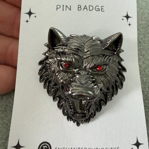 May include: A gunmetal-coloured wolf head pin badge with bright red eyes, mounted on a white display card featuring the text PIN BADGE and ENCHANTEDCURIOSITYS, decorated with small black star icons in the corners of the card.