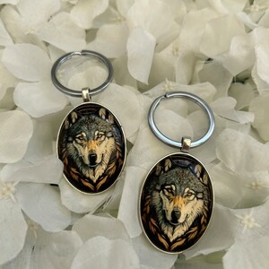 May include: Two silver keyrings with oval-shaped charms featuring a wolf's face with a brown, black, and white fur pattern. The wolf's face is surrounded by a brown and gold design.