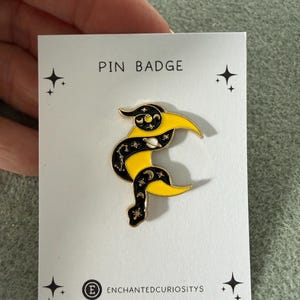 May include: A gold-toned metal pin badge on a white backing card labelled PIN BADGE and ENCHANTEDCURIOSITYS. The pin features a crescent moon shape with a black and yellow enamel design, decorated with small white stars, a planet, and moon phases.