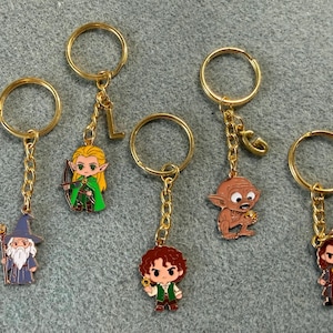 May include: Five gold-toned metal keychains featuring enamel character charms. The charms depict a wizard with a staff, an elf with a bow, a curly-haired figure, a creature holding a ring, and a warrior with a sword. Two keychains include letter charms.