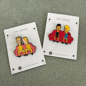 May include: Two enamel pin badges on white backing cards labelled PIN BADGE and ENCHANTEDCURIOSITYS. One pin features two characters in yellow hazmat suits on a pink couch. The other pin shows two characters in casual clothes on a red couch.