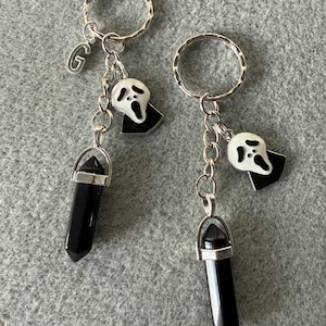 May include: Two keyrings with silver rings, each featuring a black crystal pendant, a white and black ghost charm, and a chain. One keyring has a silver letter "G" charm.