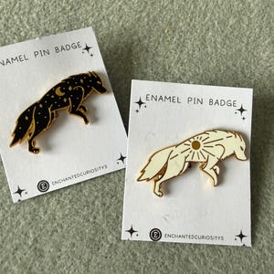 May include: Two enamel pin badges with a wolf design. One is black with a celestial pattern, the other is cream with a sun design. Each pin is on a white card with the text "ENAMEL PIN BADGE" and the shop name.