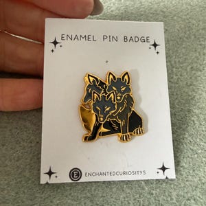May include: An enamel pin badge with a gold outline of three wolves in a stacked design. The wolves are black with grey details. The pin is on a white card with the text "ENAMEL PIN BADGE" and the seller's logo.