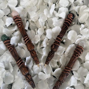May include: Four wooden pens with different coloured gemstone tops. The pens are wrapped with copper wire and have a wood grain pattern. The gemstones are green, pink and black.