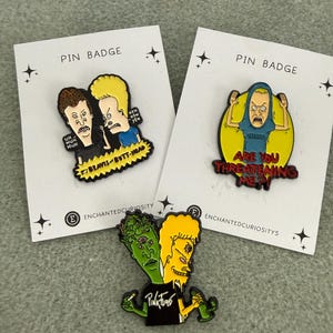 May include: Three enamel pin badges featuring Beavis and Butt-Head characters. One pin shows the duo with text "BEAVIS AND BUTT-HEAD". Another shows a character in a Metallica shirt with "ARE YOU THREATENING ME?". The third shows a two-headed figure.