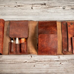 May include: A brown leather roll-up case with multiple pockets and slots for holding tools or supplies. The case is open and laid flat on a wooden surface.