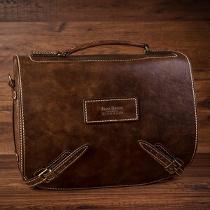 May include: A brown leather satchel bag with a top handle and adjustable shoulder strap. The bag has a stitched design and a small leather label with the text "Casa Bruno Handmade" on the front.