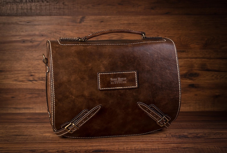 May include: A brown leather satchel bag with a top handle and adjustable shoulder strap. The bag has a stitched design and a small leather label with the text "Casa Bruno Handmade" on the front.