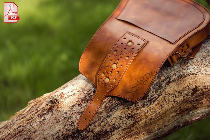 Puede incluir: Un bolso de cuero marr&oacute;n con cierre de solapa y una correa decorativa con un patr&oacute;n de c&iacute;rculos. El bolso tiene una hebilla de metal y un lazo de metal en la correa. El bolso est&aacute; sobre una superficie de madera.