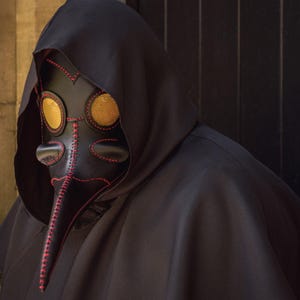 May include: A black leather plague doctor mask with a long beak and two round yellow eye pieces. The mask is decorated with red stitching.
