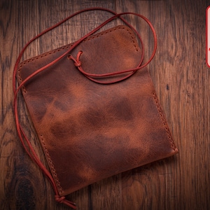 May include: A brown leather pouch with a red drawstring closure. The pouch is lying on a wooden surface.