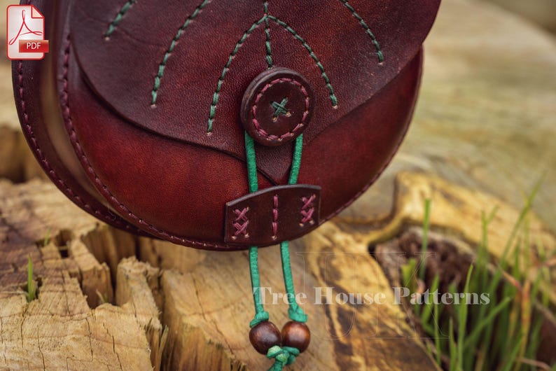 May include: A burgundy leather bag with green stitching and a green cord with wooden beads. The bag has a flap with a decorative button and a small pocket on the front.