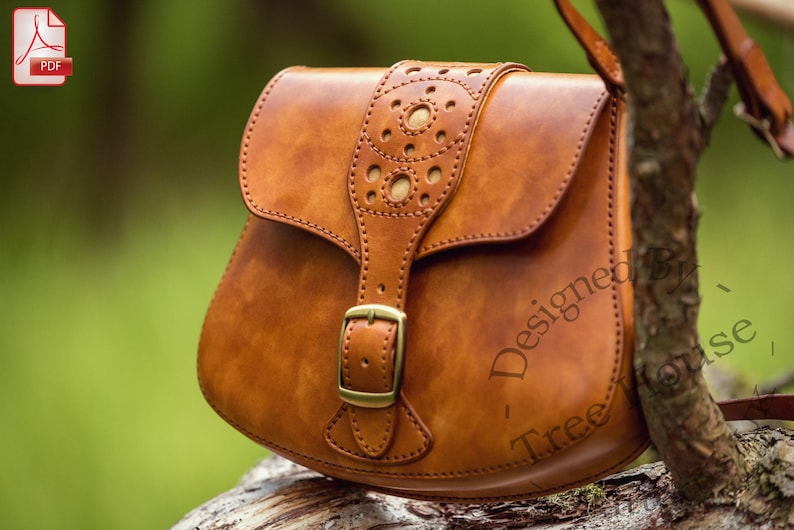 Puede incluir: Un bolso de cuero marr&oacute;n cruzado con cierre de hebilla y una correa decorativa con recortes. El bolso est&aacute; sentado en una rama de &aacute;rbol.