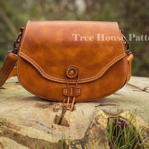 Peut inclure: Un sac bandoulière en cuir marron clair avec un rabat incurvé et des coutures blanches. Le sac est fermé par un bouton et possède une longue bandoulière réglable. Le texte "Tree House Patterns" est visible.