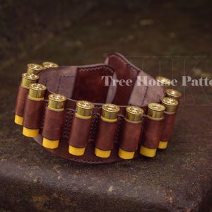 May include: A brown leather shotgun shell holder with 12 yellow shotgun shells. The holder is on a brown surface. The text 'Tree House Patterns' is visible on the holder.