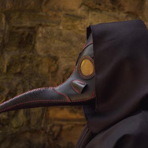 May include: A black leather plague doctor mask with a long, curved beak and a gold-colored eye piece. The mask is worn by a person in a black hooded robe.