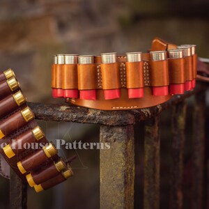 May include: Two leather shotgun shell belts, one brown and one dark brown, with rows of shotgun shells. The belts are displayed on a rusty metal fence. The text 'Tree House Patterns' is visible on the bottom of the image.