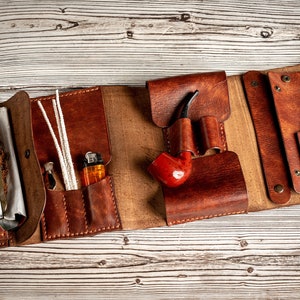 May include: A brown leather tobacco pouch with multiple compartments. The pouch is open and shows a pack of tobacco, a lighter, pipe cleaners, and a pipe.