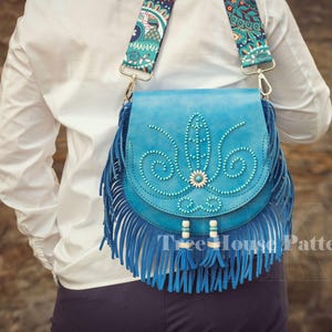 Op de afbeelding: Een blauwe leren schoudertas met franjes en een band met een bloemenpatroon. De tas heeft een parelmoerontwerp en is versierd met turquoise accenten.