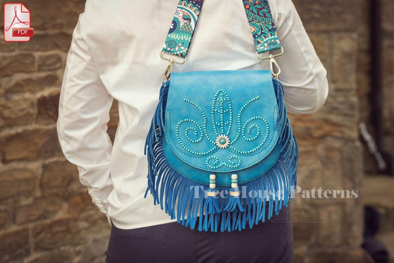 Op de afbeelding: Een blauwe leren schoudertas met franjes en een band met een bloemenpatroon. De tas heeft een parelmoerontwerp en is versierd met turquoise accenten.