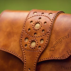 Puede incluir: Un bolso de cuero marr&oacute;n con una solapa decorativa que presenta un patr&oacute;n circular y costuras. El bolso est&aacute; estampado con el texto "Designed By...Free House Patterns".