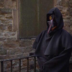 May include: A person wearing a black hooded robe and a plague doctor mask with a long beak. The mask is made of leather and has a red and black design. The person is standing in front of a stone wall.