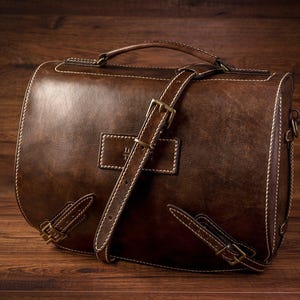May include: A brown leather briefcase with a long strap and a buckle closure. The bag has a stitched design and a flap with a metal clasp.