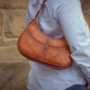 Op de afbeelding: Een bruine leren schoudertas met een gebogen vorm en zichtbare stiksels. De tas heeft een riem en wordt over de schouder gedragen. De tekst "Designed by House Patterns" is zichtbaar op de tas.