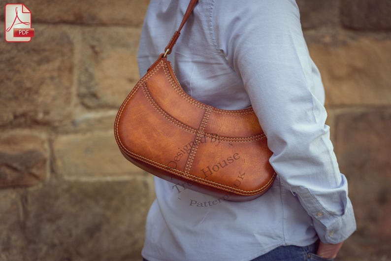 Op de afbeelding: Een bruine leren schoudertas met een gebogen vorm en zichtbare stiksels. De tas heeft een riem en wordt over de schouder gedragen. De tekst "Designed by House Patterns" is zichtbaar op de tas.