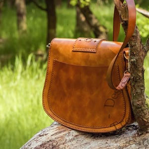 Puede incluir: Un bolso de cuero marr&oacute;n con cierre de solapa y una correa larga. El bolso tiene un dise&ntilde;o cosido y est&aacute; sentado en un tronco de &aacute;rbol.