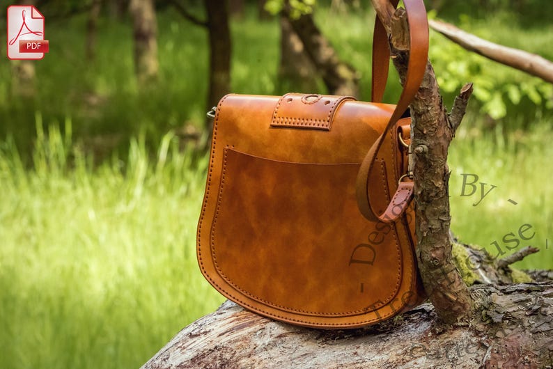 Puede incluir: Un bolso de cuero marr&oacute;n con cierre de solapa y una correa larga. El bolso tiene un dise&ntilde;o cosido y est&aacute; sentado en un tronco de &aacute;rbol.