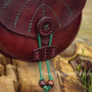 May include: A close-up of a burgundy leather bag with green stitching and a green cord with wooden beads. The bag has a flap with a button closure and a decorative design on the front. The text "Tree House Patterns" is visible in the bottom right corner of the image.