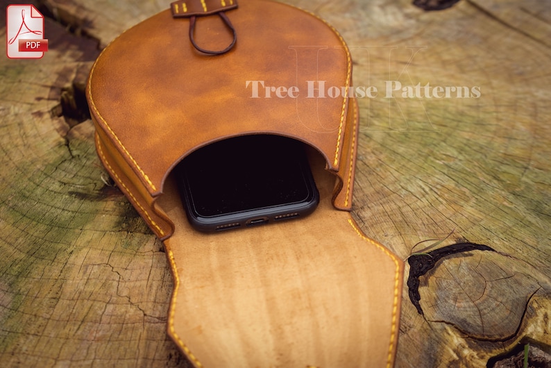 May include: A brown leather phone pouch with a flap that opens to reveal a black smartphone inside. The pouch has yellow stitching and a small loop at the top. The pouch is sitting on a wooden surface.