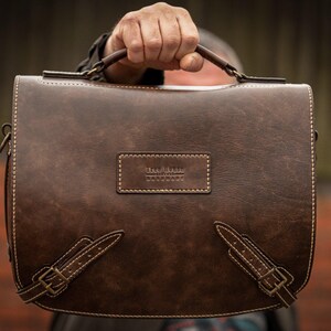 May include: A brown leather briefcase with a top handle and a buckle closure. The briefcase has a small label on the front that reads "The Leather Goods Co."
